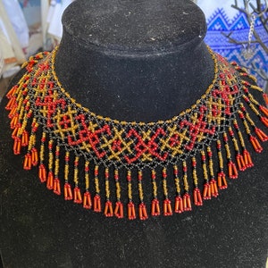 May include: A beaded necklace with a geometric pattern in red, gold, and black. The necklace has a fringe of beads hanging from the bottom.