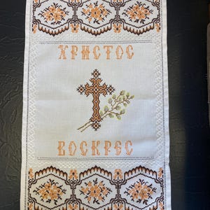 May include: White embroidered fabric with a cross, a branch of leaves, and the words "ХРИСТОС ВОСКРЕС" in orange thread. The fabric has an orange fringe border with a floral pattern.