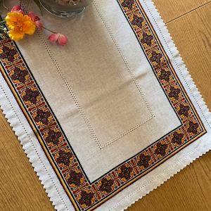 May include: A white linen table runner with a scalloped edge and a colorful embroidered border. The border features geometric patterns in shades of red, yellow, green, and black.