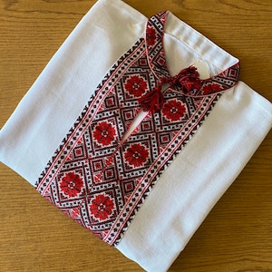 May include: White linen shirt with a red and black embroidered design on the collar. The embroidery features a geometric pattern with red flowers.