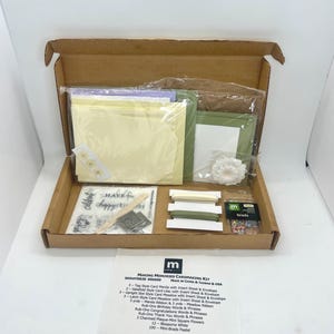 May include: Open cardboard box containing a cardmaking kit. The kit includes envelopes, paper, ribbon, embellishments, and a sheet with text that reads "MAKING MEMORIES CARDMAKING KIT". The kit is designed for creating handmade cards.