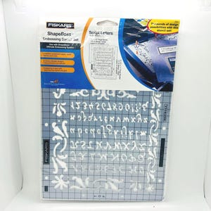 May include: A Fiskars ShapeBoss embossing stencil set featuring script letters and decorative flourishes on a clear grid sheet. The packaging includes blue and white branding with text detailing the use of the stencil for scrapbook pages.