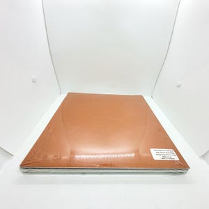 May include: A rectangular stack of flat items encased in clear plastic shrink wrap. The top surface is a solid copper-orange color. A small white rectangular sticker is affixed to the bottom right corner with the text SUFFOCATION WARNING.