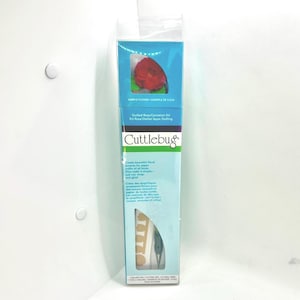 May include: A Cuttlebug Quilled Rose/Carnation Kit in a tall, rectangular box. The packaging features a blue and white design with a red paper flower sample at the top and a quilling tool visible through a clear window at the bottom.