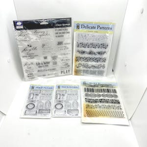 May include: A collection of clear stamps in plastic packaging. The stamps feature various designs, including inspirational quotes, decorative borders, and label templates. The text "Clear Stamps" and "Delicate Patterns" are visible.