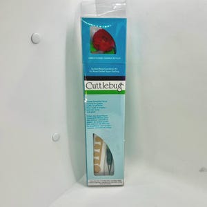 May include: A Cuttlebug Quilled Rose/Carnation Kit in a tall, rectangular clear plastic box. The packaging features a red paper flower sample at the top, the Cuttlebug brand logo, and text describing the kit for creating floral paper crafts.