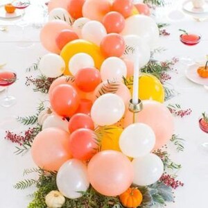Pode incluir: Um centro de mesa feito com uma guirlanda de bal&otilde;es laranja, amarelo e branco com vegeta&ccedil;&atilde;o e pequenas ab&oacute;boras.