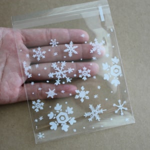Puede incluir: Una bolsa de pl&aacute;stico transparente con cierre de cremallera, decorada con dise&ntilde;os de copos de nieve blancos de varios tama&ntilde;os. La bolsa est&aacute; sostenida por una mano, mostrando el dise&ntilde;o. Los copos de nieve est&aacute;n esparcidos por la superficie, creando un tema invernal.
