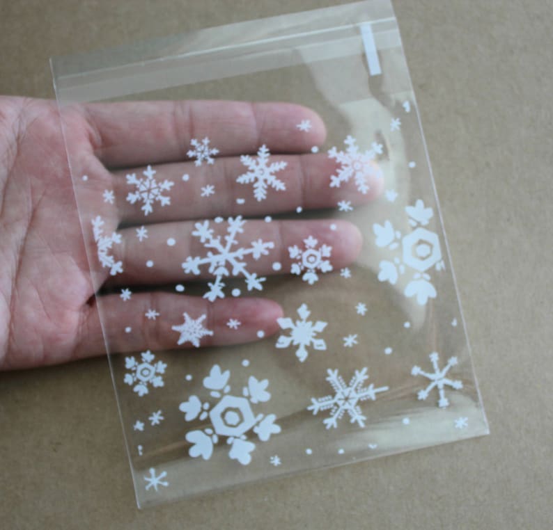 Puede incluir: Una bolsa de pl&aacute;stico transparente con cierre de cremallera, decorada con dise&ntilde;os de copos de nieve blancos de varios tama&ntilde;os. La bolsa est&aacute; sostenida por una mano, mostrando el dise&ntilde;o. Los copos de nieve est&aacute;n esparcidos por la superficie, creando un tema invernal.