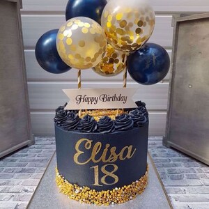 Pode incluir: Um bolo de anivers&aacute;rio preto e dourado com as palavras "Happy Birthday" e "Elisa 18" nele. O bolo &eacute; decorado com confeitos dourados e coberto com bal&otilde;es azuis e dourados.