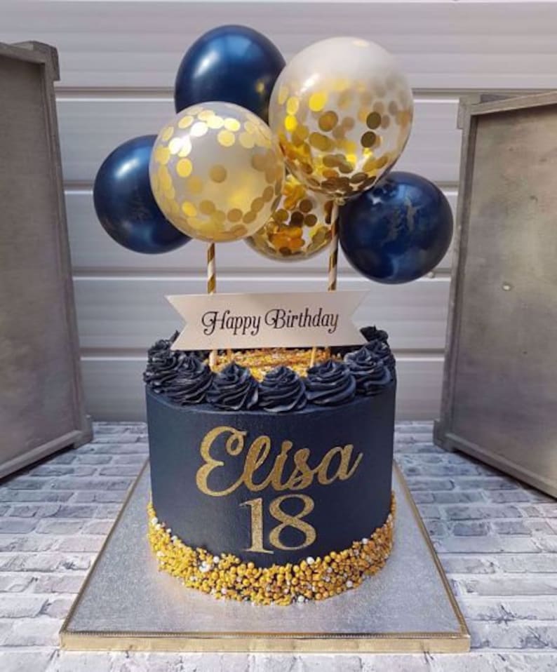 Pode incluir: Um bolo de anivers&aacute;rio preto e dourado com as palavras "Happy Birthday" e "Elisa 18" nele. O bolo &eacute; decorado com confeitos dourados e coberto com bal&otilde;es azuis e dourados.