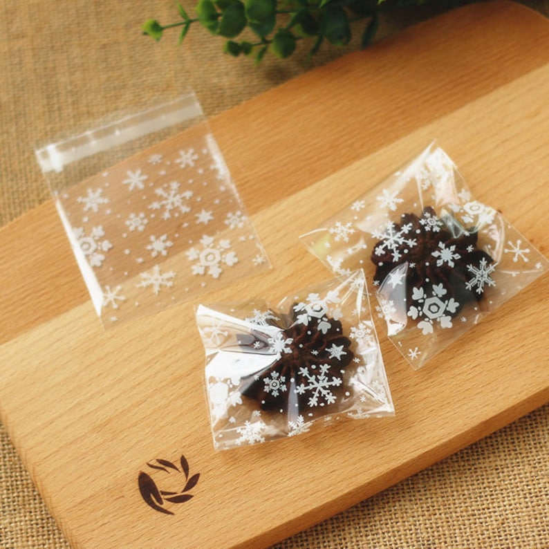 Puede incluir: Bolsas transparentes de celof&aacute;n con dise&ntilde;os de copos de nieve blancos, que contienen galletas de chocolate en forma de estrella. Las bolsas est&aacute;n selladas y dispuestas sobre una tabla de cortar de madera, evocando un tema invernal.