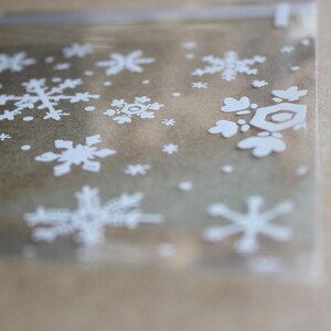 Puede incluir: Una bolsa de celof&aacute;n transparente y rectangular con un patr&oacute;n repetido de copos de nieve blancos. Los copos de nieve var&iacute;an en tama&ntilde;o y dise&ntilde;o, creando un tema de vacaciones de invierno. La bolsa es parcialmente transparente.