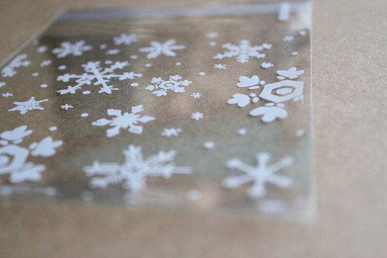 Puede incluir: Una bolsa de celof&aacute;n transparente y rectangular con un patr&oacute;n repetido de copos de nieve blancos. Los copos de nieve var&iacute;an en tama&ntilde;o y dise&ntilde;o, creando un tema de vacaciones de invierno. La bolsa es parcialmente transparente.