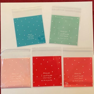 May include: Five small, clear plastic bags with colorful backgrounds. The bags have a teal, green, pink, and two red backgrounds, each with white polka dots. Each bag has the text "Because I want to see your face with a smile."