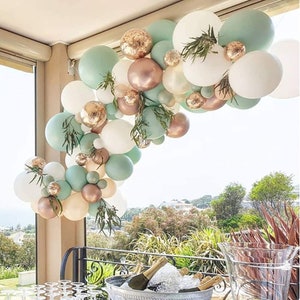 May include: A balloon arch made of white, sage green, and rose gold balloons. The arch is decorated with greenery and is perfect for a wedding, party, or other special event.