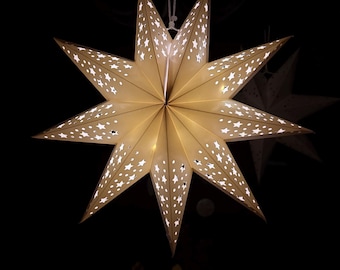 May include: A seven-pointed star-shaped paper lantern in a warm off-white color, featuring numerous small star-shaped cutouts that allow light to shine through. The lantern is suspended from a white cord against a dark, solid black background.
