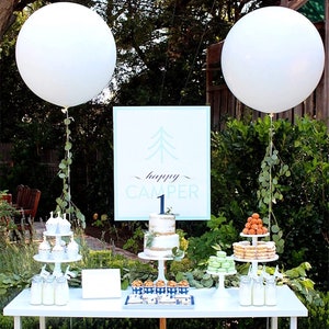 May include: A white table with a dessert table setup for a first birthday party. The table is decorated with white balloons, greenery, and a sign that says "happy camper 1". There are cupcakes, cookies, macarons, and a cake on the table.