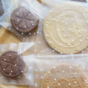 May include: Clear plastic bags with white polka dots, perfect for packaging small treats like cookies. The bags are shown with two different types of cookies, one with a brown and white design and the other with a light brown and white design.