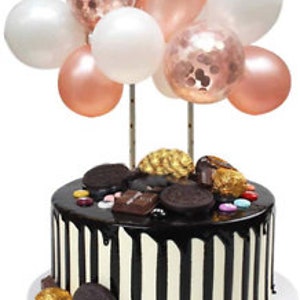 Pode incluir: Um bolo de chocolate com bal&otilde;es brancos e rosa dourado no topo. O bolo &eacute; decorado com raspas de chocolate, doces e um fio de chocolate.