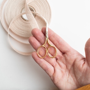 May include: A pair of small gold-coloured scissors with silver blades, held in a hand against a white background.