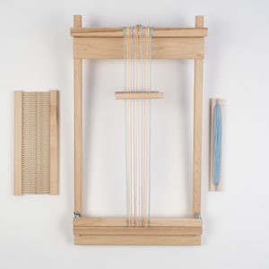 May include: A wooden loom with a set of colorful threads strung across it. The loom is made of natural wood and has a simple design. There is a separate wooden shuttle and a skein of blue yarn.