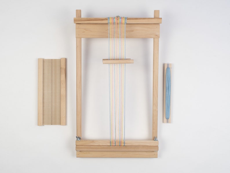 May include: A wooden loom with a set of colorful threads strung across it. The loom is made of natural wood and has a simple design. There is a separate wooden shuttle and a skein of blue yarn.