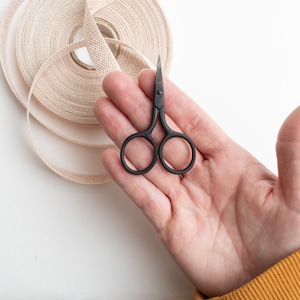 May include: A pair of small, black scissors rests in an open hand, next to a roll of beige ribbon. The scissors have a classic design, and the ribbon is neatly coiled. The background is white.