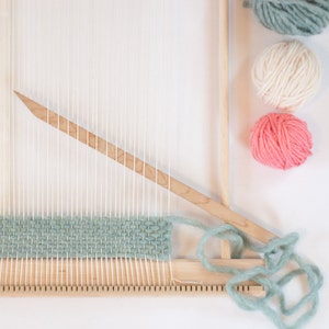 Peut inclure: Un m&eacute;tier &agrave; tisser en bois avec du fil vert, blanc et rose. Le m&eacute;tier &agrave; tisser est install&eacute; avec une navette et une aiguille &agrave; tisser. Le fil est enroul&eacute; autour du m&eacute;tier &agrave; tisser et pr&ecirc;t &agrave; &ecirc;tre tiss&eacute;.