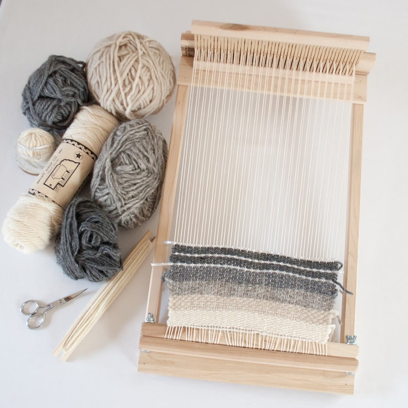 May include: A wooden loom with a partially woven tapestry in shades of gray and beige.  The loom is surrounded by balls of yarn in various shades of gray and beige.
