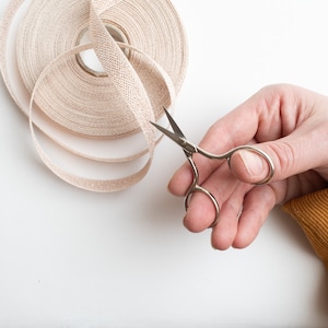May include: A hand holding a pair of silver scissors cutting a roll of beige ribbon.