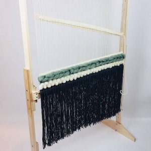 May include: A wooden loom with a black and white woven tapestry with a green fringe. The loom is freestanding and has a wooden base.