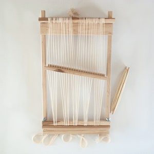 May include: A wooden loom with white string warp threads and a wooden shuttle. The loom is set up for weaving and has a white background.
