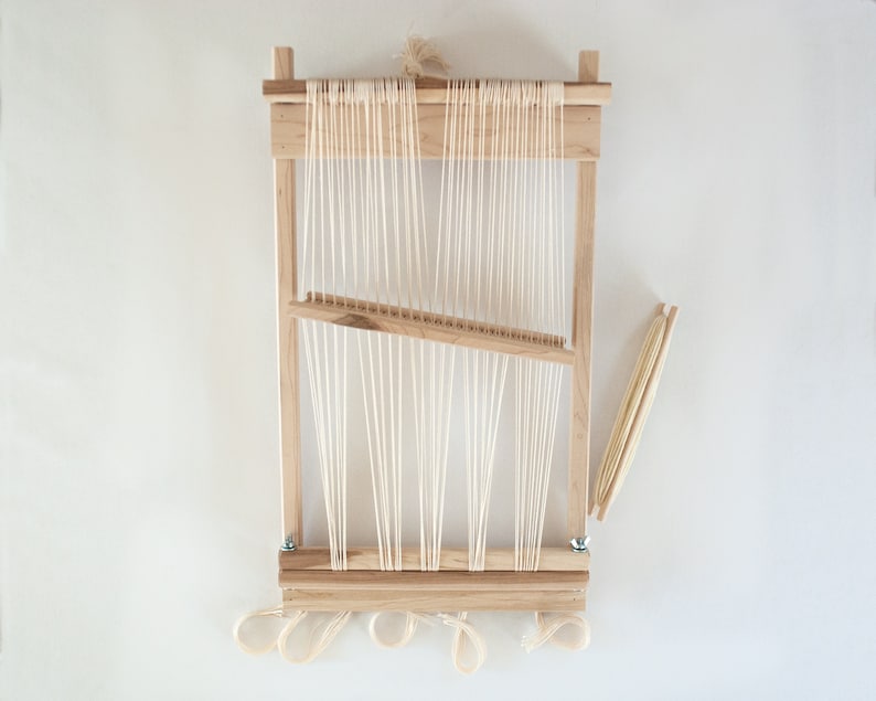 May include: A wooden loom with white string warp threads and a wooden shuttle. The loom is set up for weaving and has a white background.