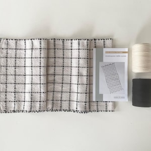 May include: A woven table runner with a black and white grid pattern and fringe. A pattern booklet and two spools of thread, one white and one black, are also included. The booklet reads "windowpane table runner".