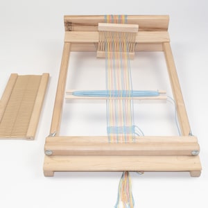 May include: A wooden loom with a blue, yellow, pink, and orange yarn warp. The loom has a wooden shuttle and a wooden heddle. The loom is set up for weaving.