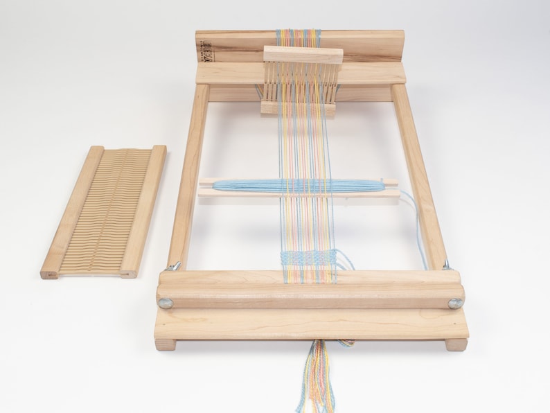 May include: A wooden loom with a blue, yellow, pink, and orange yarn warp. The loom has a wooden shuttle and a wooden heddle. The loom is set up for weaving.