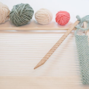 Peut inclure: Un m&eacute;tier &agrave; tisser en bois avec un fil bleu clair tiss&eacute; dans les fils de cha&icirc;ne. Quatre pelotes de fil sont en arri&egrave;re-plan : une blanche, une bleu clair, une rose et une blanche.