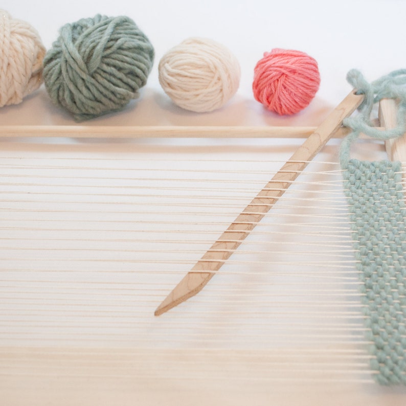Peut inclure: Un m&eacute;tier &agrave; tisser en bois avec un fil bleu clair tiss&eacute; dans les fils de cha&icirc;ne. Quatre pelotes de fil sont en arri&egrave;re-plan : une blanche, une bleu clair, une rose et une blanche.