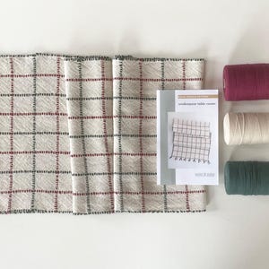 May include: A woven table runner with a cream-colored background and a grid pattern in red and black. The runner has fringed edges with red and green accents. Three spools of thread in burgundy, cream, and green are next to a pattern.