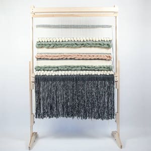May include: A wooden loom with a woven tapestry featuring a variety of colors and textures, including green, pink, and white. The tapestry has a fringe at the bottom.