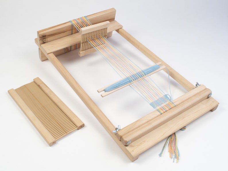 May include: A wooden loom with a blue, yellow, and pink yarn threaded through it. The loom is set up for weaving and has a small wooden shuttle. The loom is on a white surface.