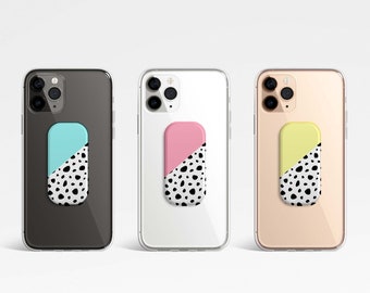 Puede incluir: Tres fundas de teléfono con agarres de teléfono de diferentes colores. Las fundas de teléfono son negras, blancas y doradas. Los agarres de teléfono son azules, rosas y amarillos. Cada agarre tiene un patrón de lunares negros y blancos.