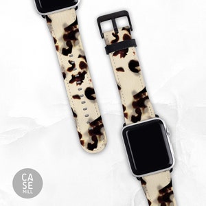 Pode incluir: Duas pulseiras de Apple Watch com fundo creme e padrão tartaruga. Uma pulseira está presa a um Apple Watch preto, e a outra está solta. As pulseiras têm ferragens pretas.