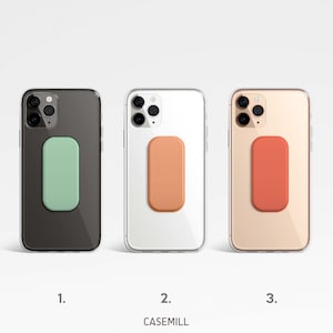 Puede incluir: Tres fundas transparentes para teléfono con agarres de teléfono de diferentes colores. Las fundas son negra, blanca y dorada. Los agarres son verde, naranja y rojo.
