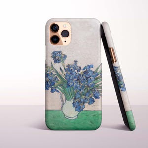 May include: A phone case displaying a painting of blue irises in a white vase. The artwork is set against a light background with a green base. Another case with a similar design is leaning against it.