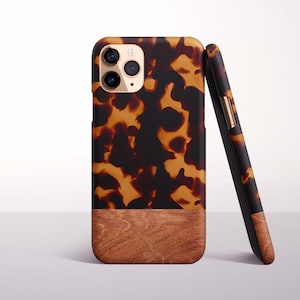 Op de afbeelding: Een bruine en zwarte telefoonhoes met schildpadpatroon en een houten nerf bodem. De telefoonhoes wordt rechtopstaand en tegen een tweede hoes geleund getoond. De telefoonhoes heeft een camera en een flitser.