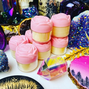 May include: Six small pink containers with a rose design on the lid. The containers are filled with a light yellow substance. The containers are arranged in a pyramid shape.