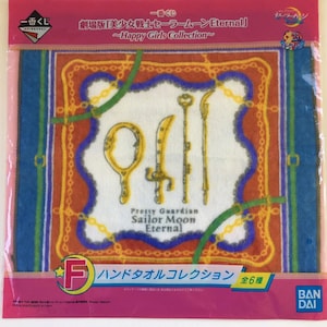 May include: A packaged Sailor Moon Eternal hand towel. The white towel features gold-colored weapon illustrations and the text "Pretty Guardian Sailor Moon Eternal." The towel is framed by a blue and orange border with gold accents. The packaging is pink and blue.