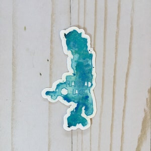 May include: A blue and green watercolor sticker of Flathead Lake. The sticker is on a white wooden background.